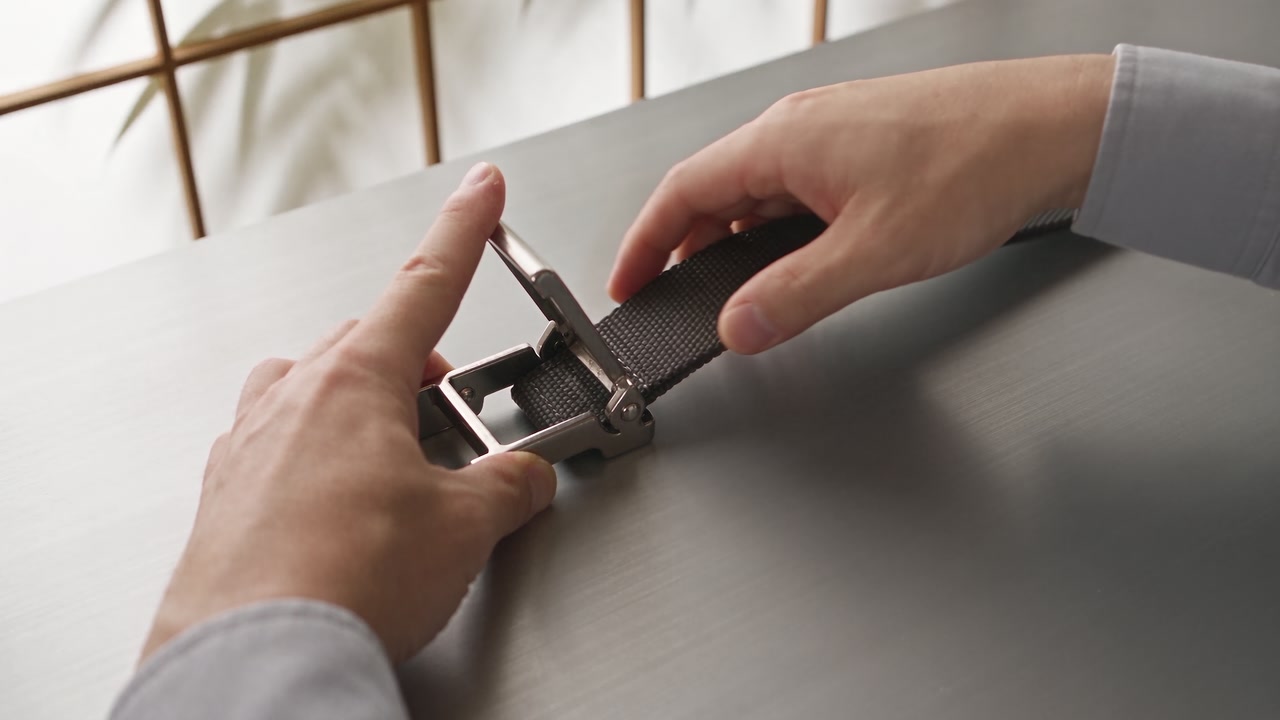 Two hands thread a fabric strap through a metal buckle on a grey desk